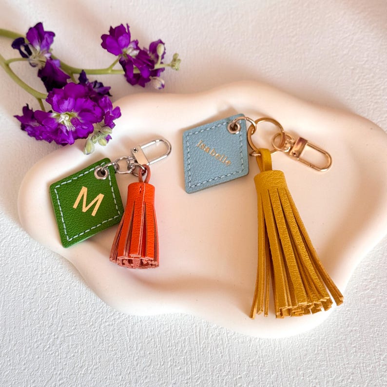 May include: Three leather keychains with tassels. The keychains are in the shape of a square with rounded corners. The tassels are orange, yellow, and green. The keychains are labeled with the letters "M" and "Isabelle".