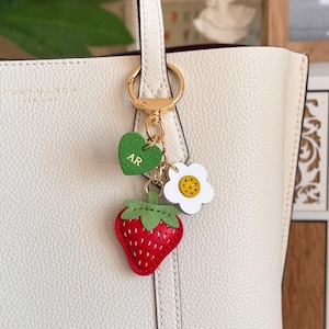 Puede incluir: Un llavero de metal dorado sujeto a un bolso de mano blanco texturizado. El llavero cuenta con tres dijes: un coraz&oacute;n verde grabado con las letras AR, una flor blanca con centro amarillo y una fresa roja con hojas verdes.