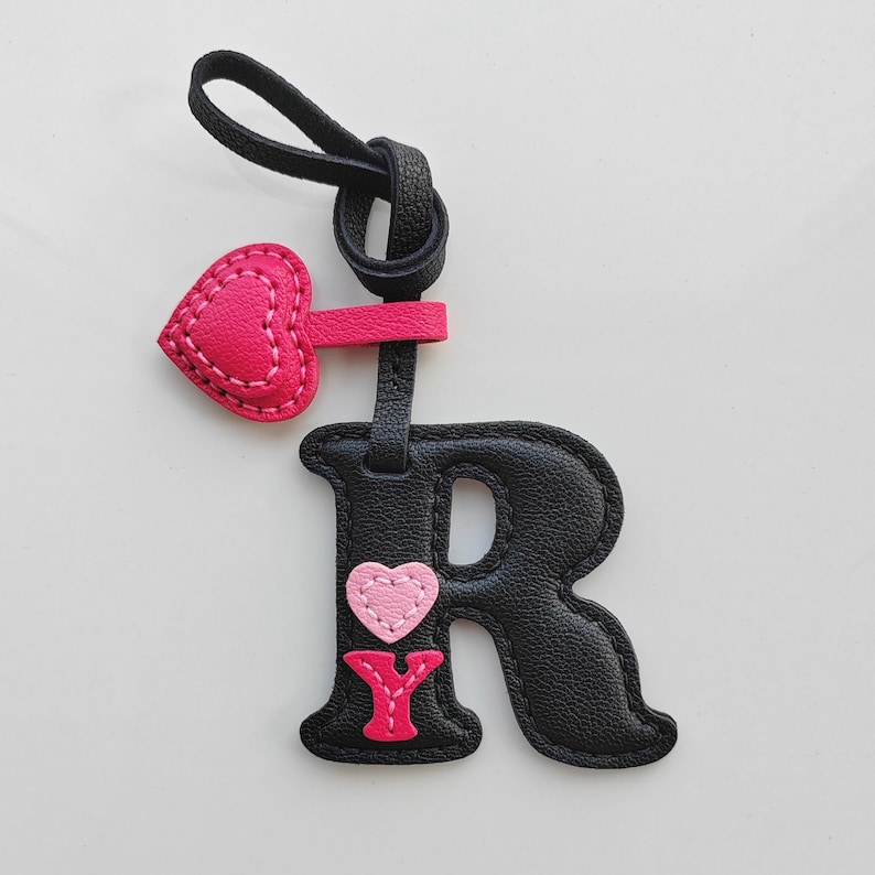 Puede incluir: Un llavero con la letra R negra con superficie texturizada y bordes cosidos. Presenta un peque&ntilde;o coraz&oacute;n rosa y una letra Y rosa brillante adheridos al frente. Un dije de coraz&oacute;n rosa intenso cuelga del lazo de la correa negra en la parte superior.