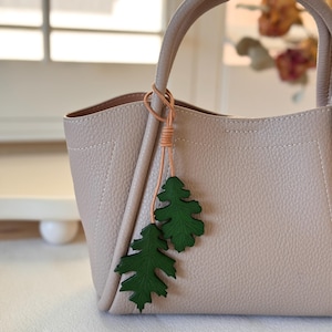 May include: A light beige leather tote bag with a brown leather strap and a green leather leaf charm.