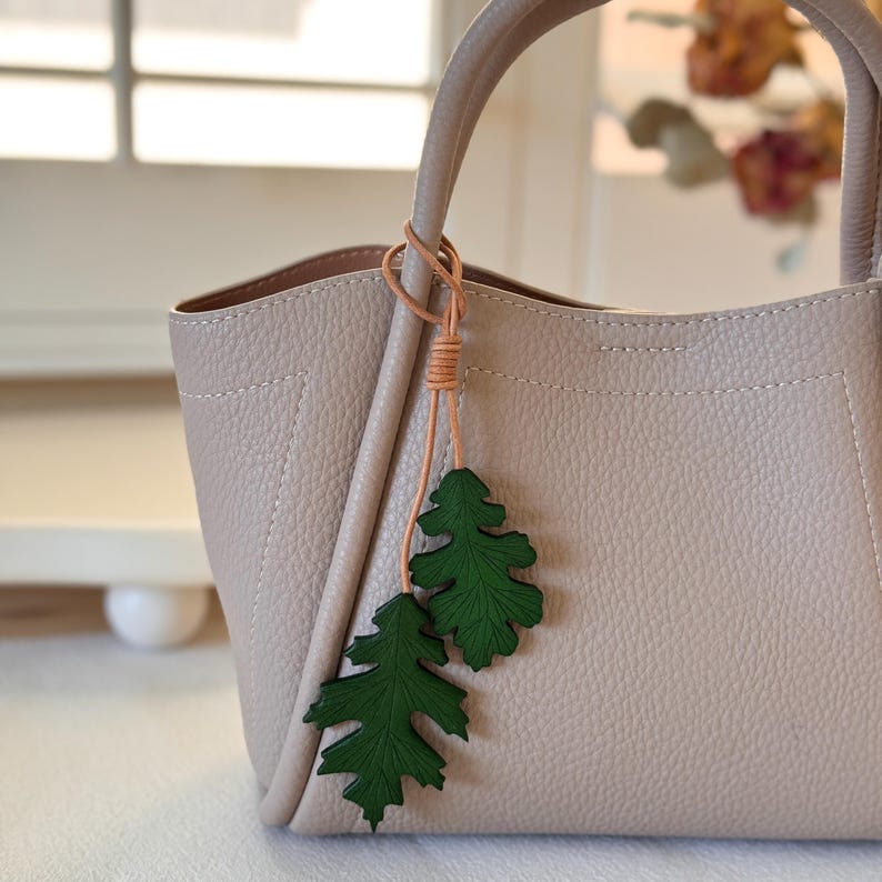 May include: A light beige leather tote bag with a brown leather strap and a green leather leaf charm.