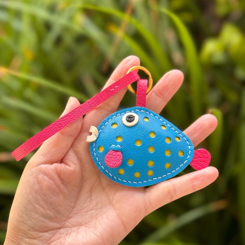 Puede incluir: Un llavero de cuero de pez globo azul y amarillo con detalles rosas y una correa rosa.