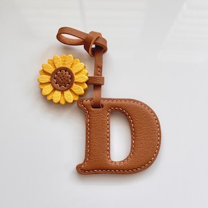 Puede incluir: Una etiqueta de equipaje de cuero marrón con la letra "D" y un adorno de girasol. El girasol es amarillo con el centro marrón. La etiqueta tiene un lazo para sujetar.