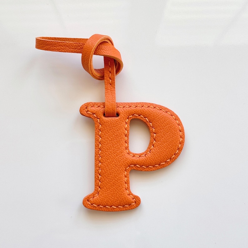 May include: Orange leather luggage tag in the shape of the letter "P". The tag has a loop at the top with a knotted detail and is stitched around the edges with a contrasting thread. The tag is made of textured leather.