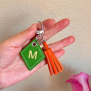 May include: Green leather keychain with a diamond-shaped tag that says "M" in gold lettering. The keychain also has an orange leather tassel.