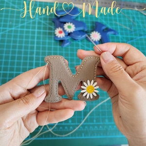 Puede incluir: Primer plano de una mano cosiendo una letra "M" de cuero marr&oacute;n con una margarita blanca. La letra se est&aacute; cosiendo sobre una estera verde con la palabra "Hand Made" en el fondo.