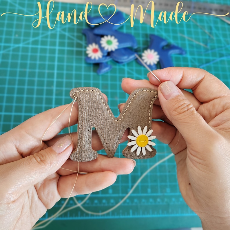 Puede incluir: Primer plano de una mano cosiendo una letra "M" de cuero marr&oacute;n con una margarita blanca. La letra se est&aacute; cosiendo sobre una estera verde con la palabra "Hand Made" en el fondo.