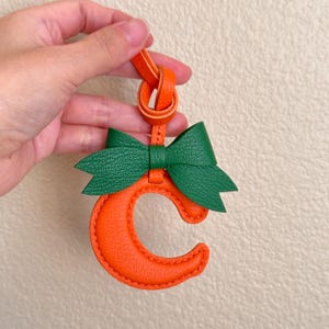 Puede incluir: Llavero de cuero naranja con un lazo de cuero verde y la letra "C" cosida con hilo naranja.