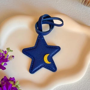 Puede incluir: Un dije de cuero azul marino en forma de estrella con superficie texturizada, costuras gruesas en azul marino a lo largo de los bordes y una pequeña aplicación de media luna amarilla. El dije está unido a una correa de cuero azul marino.