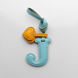 Puede incluir: Un llavero con la letra J de color azul claro texturizado, con una correa de lazo a juego y un peque&ntilde;o dije de coraz&oacute;n amarillo unido por una banda horizontal corta. La pieza presenta costuras visibles en los bordes de la letra y el coraz&oacute;n.