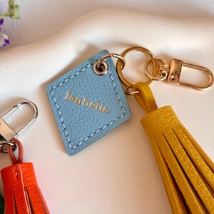 May include: A light blue leather keychain with the name "Isabelle" embossed in gold lettering. The keychain has a gold metal ring and a yellow tassel.
