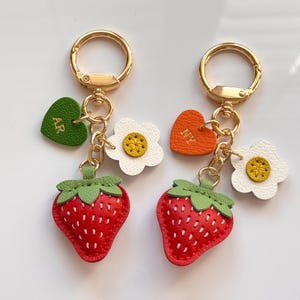 Op de afbeelding: Twee sleutelhangers met gouden ringen, elk met een rode aardbei-bedel met groene bladeren. Eén sleutelhanger heeft een groen hart met "AR", de andere een oranje hart met "NY". Beide hebben witte bloemvormige bedels.