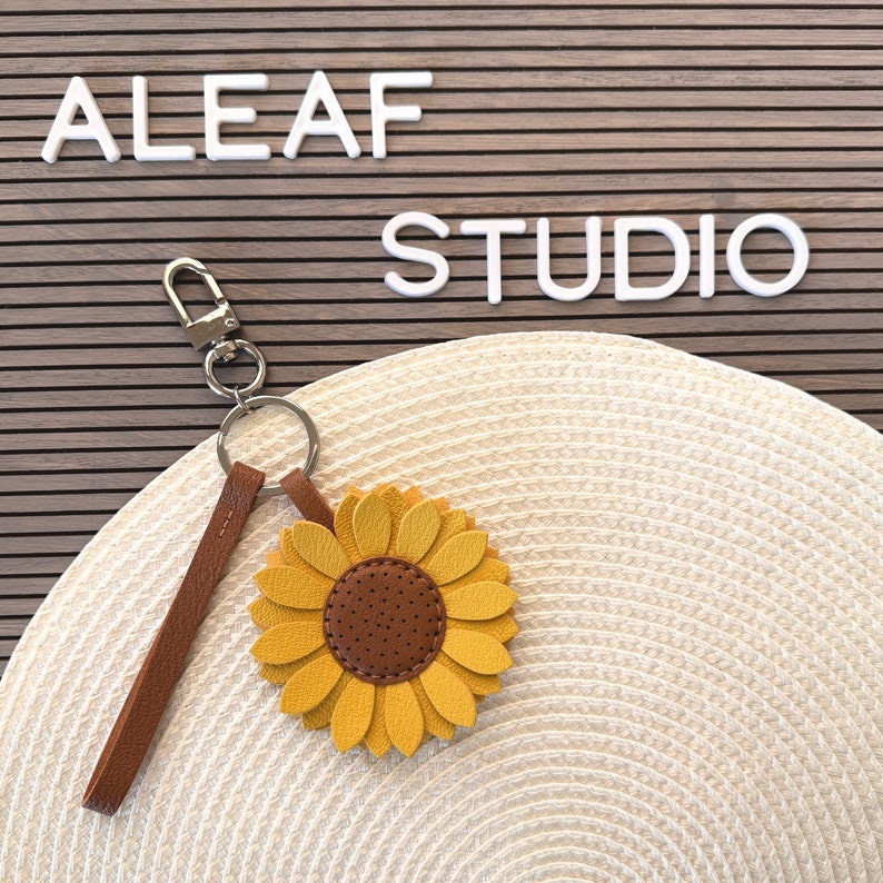May include: A brown leather keychain with a sunflower charm. The sunflower is yellow with a brown center and has a textured leather finish. The keychain has a silver metal ring and a lobster clasp.