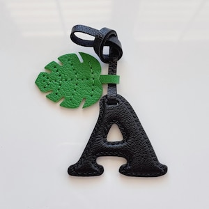 May include: Black leather letter "A" charm with a green leaf accent. The charm has a knotted loop for attachment. The leaf is a vibrant green with stitched details. A stylish accessory for bags or keychains.