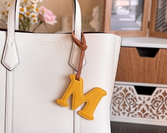 May include: White leather tote bag with a brown leather handle and a yellow letter "M" charm. The bag has a structured design with a top handle and a contrasting interior. The charm is attached with a brown leather strap.