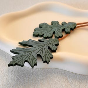 May include: Two green leather leaves with detailed vein patterns, connected by a brown cord. The leaves are on a white surface.