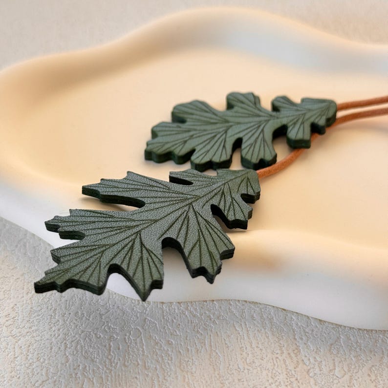 May include: Two green leather leaves with detailed vein patterns, connected by a brown cord. The leaves are on a white surface.