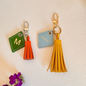 May include: Two leather keychains with tassels. One keychain is green with the letter "M" and an orange tassel. The other keychain is light blue with the name "Isabelle" and a yellow tassel.