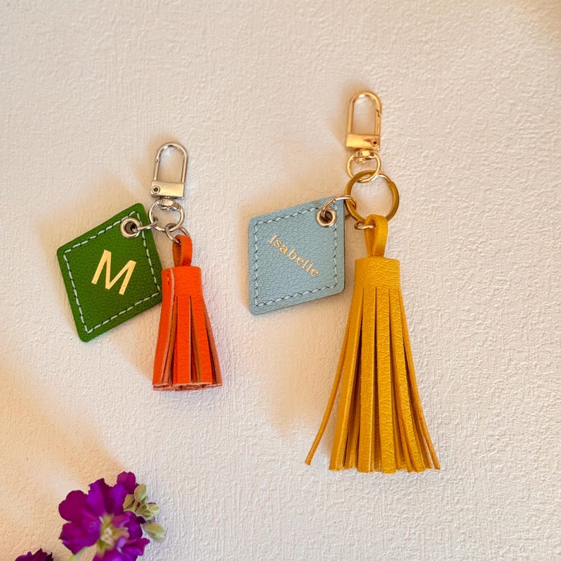 May include: Two leather keychains with tassels. One keychain is green with the letter "M" and an orange tassel. The other keychain is light blue with the name "Isabelle" and a yellow tassel.