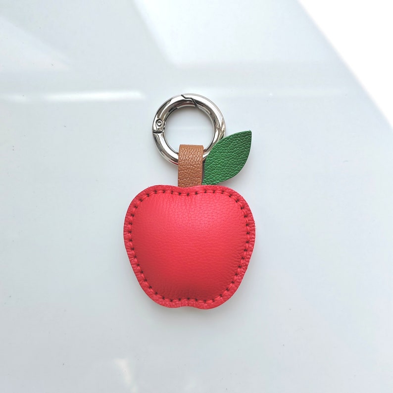 Puede incluir: Un llavero de cuero rojo en forma de manzana con una hoja de cuero verde y un anillo de llave de metal plateado.