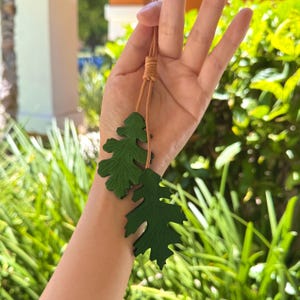 May include: A green leather necklace with two leaf charms. The necklace is made of brown leather cord and has a simple knot closure.