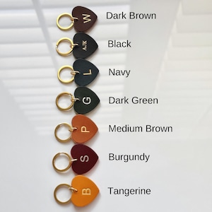 May include: Heart-shaped leather keychains in various colors: dark brown, black, navy, dark green, medium brown, burgundy, and tangerine. Each keychain has a gold-colored ring and a single initial. The colors are labeled with text.