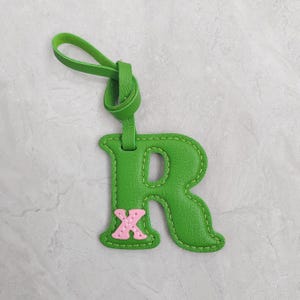 Puede incluir: Un llavero con la letra R de color verde brillante con una superficie texturizada y costuras visibles en los bordes. Una peque&ntilde;a letra X de color rosa claro est&aacute; cosida en la parte inferior izquierda de la R. Un lazo verde a juego est&aacute; arriba.