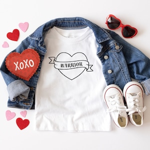 May include: White t-shirt with a black outline of a heart with the word "WARRIOR" inside the heart and a banner underneath. The shirt is laid out on a white surface with a denim jacket, red heart-shaped decorations, and white sneakers.