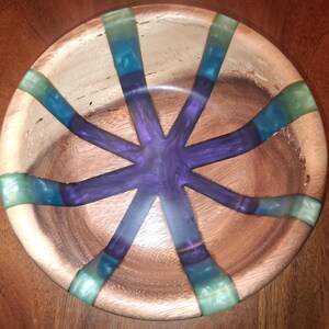 May include: A wooden bowl with a unique design. The bowl features a starburst pattern of teal, blue, and purple resin inlaid into the wood. The bowl is round and has a natural wood grain finish, creating a striking contrast with the colorful resin.
