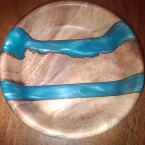 May include: A round wooden bowl with a natural wood grain finish. The bowl features three bands of turquoise resin that flow across the surface, creating a river-like design. The bowl is likely handmade.