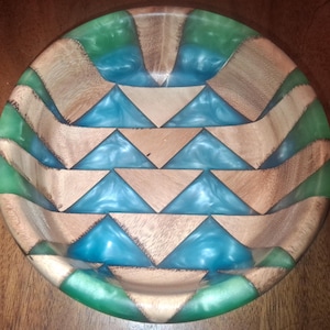 May include: A handcrafted wooden bowl with a geometric design. The bowl features alternating sections of natural wood, turquoise resin, and green resin. The bowl's interior is smooth and the exterior is textured.