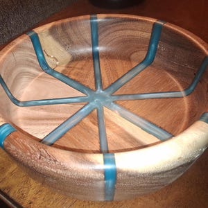 May include: A handcrafted wooden bowl with a unique design. The bowl features a natural wood grain with teal-colored resin accents that form a starburst pattern in the center. The bowl's rim also has teal resin inlays.
