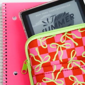 May include: A zippered tablet sleeve featuring a pink and orange checkered pattern with lime green bow illustrations, containing a tablet displaying the text The Summer, positioned against a bright pink spiral-bound notebook with a white background.