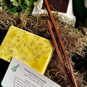 May include: A yellow Oatmeal Turmeric Acne Bar made with honey, oats, night owl oil, goat milk, and turmeric. The bar is accompanied by a card with product information and two brown sticks. The bar is surrounded by dried plants.