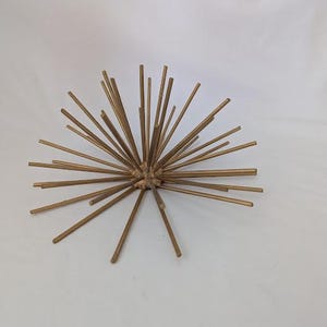 May include: A gold-colored, starburst-shaped decorative object. The sculpture features numerous slender rods radiating outwards from a central point. The rods are of varying lengths, creating a dynamic and textured appearance. The object is set against a plain white background.
