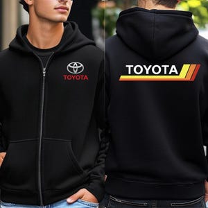May include: Black zip-up hoodie featuring the Toyota logo on the front left chest and the word "TOYOTA" with a retro stripe design on the back. The hoodie has a hood and two front pockets.