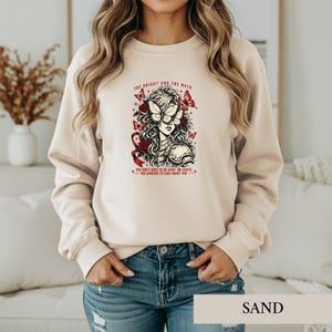 May include: A sand-colored crewneck sweatshirt featuring a graphic print of a person with a butterfly over their eyes, surrounded by red roses and butterflies. The text reads: THE BRIGHT AND THE MOTH YOU DON'T HAVE TO BE GOOD OR USEFUL.