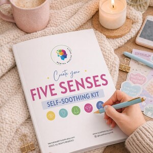 May include: A self-soothing kit titled "Create Your Five Senses" with colorful icons and text. A person is holding a light blue pen with gold accents, writing on the kit. The kit is on a cream-colored knitted blanket, with a candle and a pink mug.