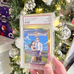 May include: A custom trading card in a clear protective slab featuring an anime-style illustration of a couple. The label reads 2025 HAPPY ANNIVERSARY MICHAEL & ELIZABETH 1ST EDITION MINT 10 I LOVE YOU. The card displays a couple standing in a field.