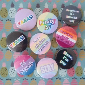 May include: A collection of round, colorful buttons with various text designs. The buttons feature phrases like "YAAAS!", "Fruity AF", "One of dem Queers", "Bi Myself", "SLAY", "Everyone is a little bit gay", "And they were roommates", and "Move, I'm gay".