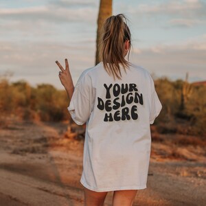 May include: A white t-shirt with the text "YOUR DESIGN HERE" in black, retro-style lettering. The shirt is being worn by a person in a desert setting.