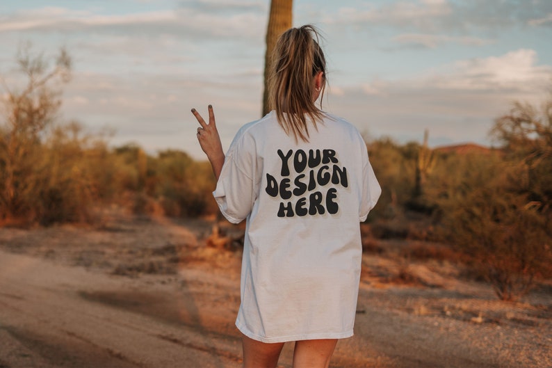 May include: A white t-shirt with the text "YOUR DESIGN HERE" in black, retro-style lettering. The shirt is being worn by a person in a desert setting.