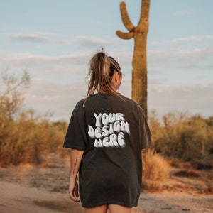 May include: A person wearing a black t-shirt with the text "YOUR DESIGN HERE" in white. The person is standing in a desert landscape with a large cactus in the background.