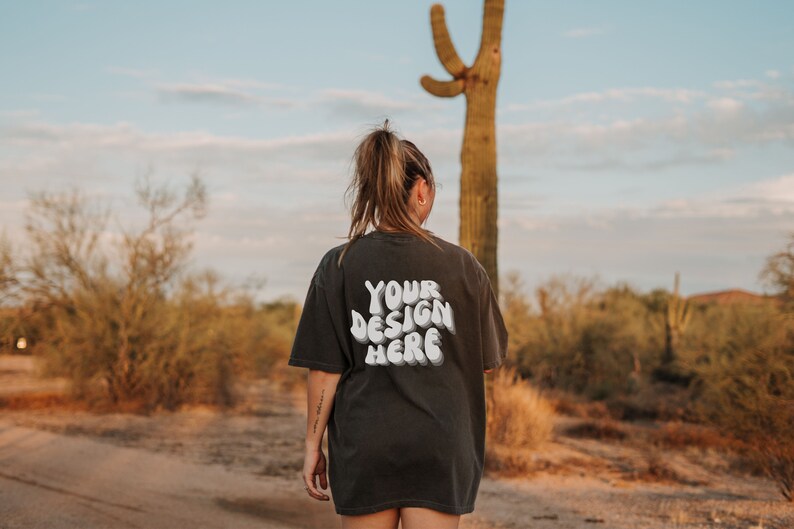 May include: A person wearing a black t-shirt with the text "YOUR DESIGN HERE" in white. The person is standing in a desert landscape with a large cactus in the background.