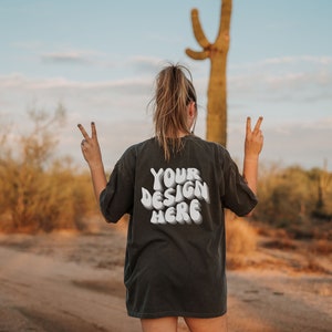 May include: A person wearing a black t-shirt with the text "YOUR DESIGN HERE" in white. The person is standing in a desert landscape with a cactus in the background.