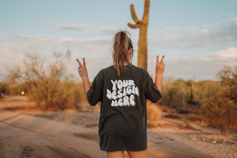 May include: A person wearing a black t-shirt with the text "YOUR DESIGN HERE" in white. The person is standing in a desert landscape with a cactus in the background.
