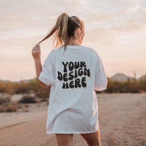 May include: A white oversized t-shirt with the text "YOUR DESIGN HERE" printed on the back in black. The shirt is being worn by a person in a desert setting.