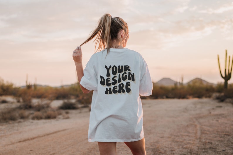 May include: A white oversized t-shirt with the text "YOUR DESIGN HERE" printed on the back in black. The shirt is being worn by a person in a desert setting.