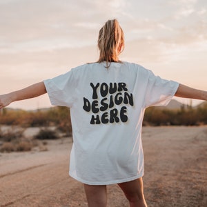 May include: A white t-shirt with the text "YOUR DESIGN HERE" in black, retro-style lettering. The shirt is being worn by a person standing in a desert landscape.