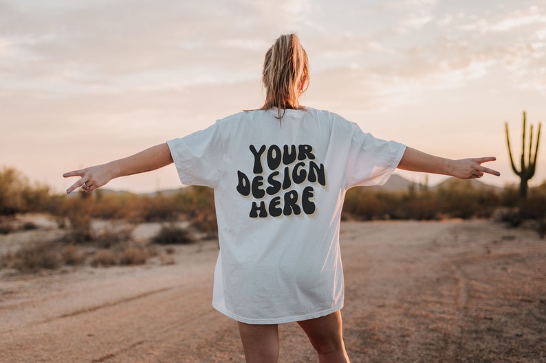 May include: A white t-shirt with the text "YOUR DESIGN HERE" in black, retro-style lettering. The shirt is being worn by a person standing in a desert landscape.
